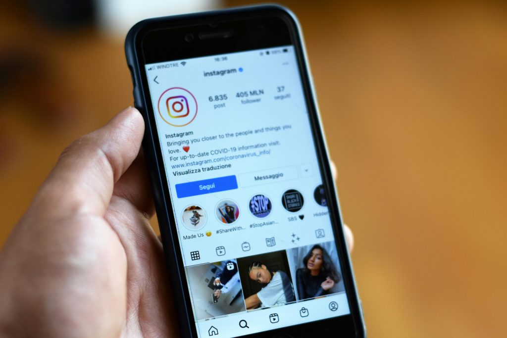 دەستێک مۆبایلی iPhone هەڵگرتووە و پرۆفایلی فەرمی Instagram نیشان دەدات لەگەڵ ژمارەی فۆڵۆوەر و بایو و ستۆریە دیارەکان